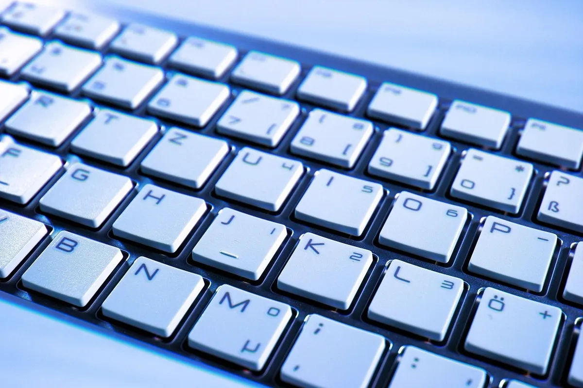 Um close-up de um teclado com teclas brancas ou muito claras, com as letras e símbolos pretos. A iluminação de fundo azul confere uma estética moderna. O layout em QWERTY é padrão, o que o torna ideal como teclado para iniciantes focando na clareza visual e conforto de digitação.