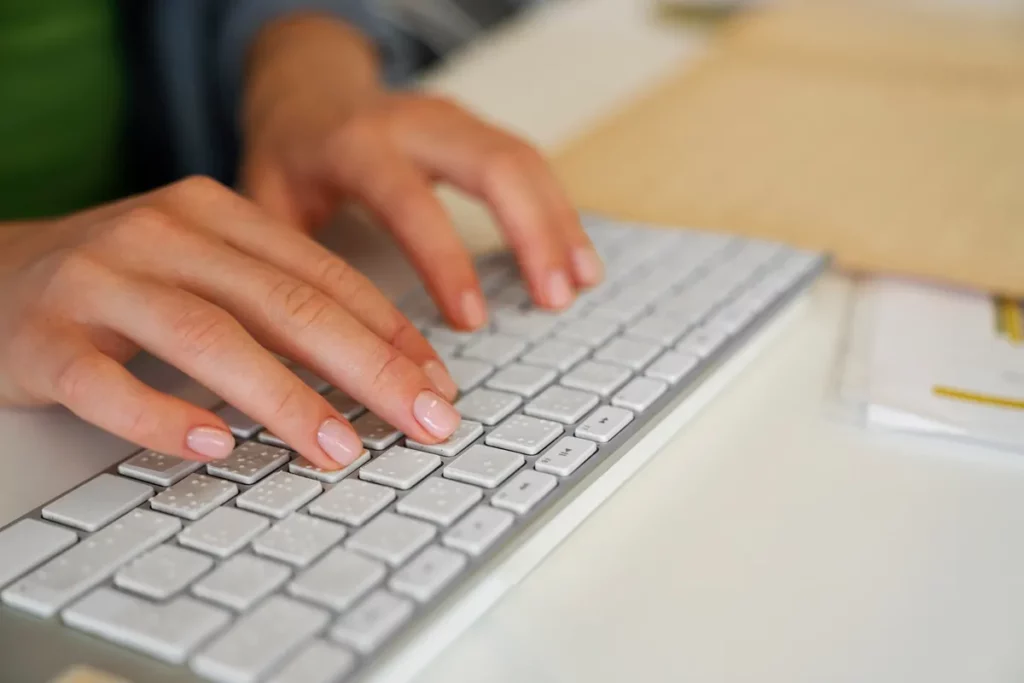 Mãos femininas digitando em um teclado branco sobre mesa de escritório organizada.