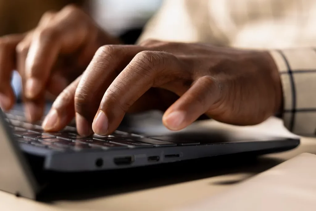 Mãos masculinas digitando em teclado de notebook preto em ambiente de escritório.