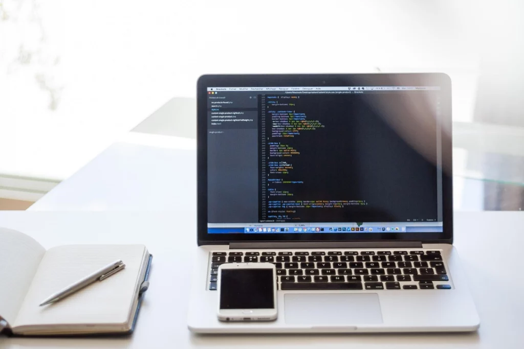 Computador em cima de uma mesa, com um celular e caderno do lado