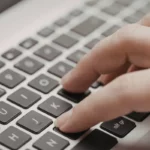 Mão digitando em um teclado de laptop prateado enquanto utiliza o computador.
