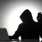 Silhueta de uma pessoa com capuz usando um laptop enquanto segura um smartphone em ambiente escuro.