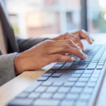 Pessoa digitando em um teclado de perfil baixo integrado a um laptop sobre uma mesa de trabalho.
