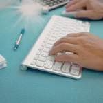 Pessoa digitando em um teclado branco sem fio sobre uma mesa azul ao lado de um mouse e um caderno.